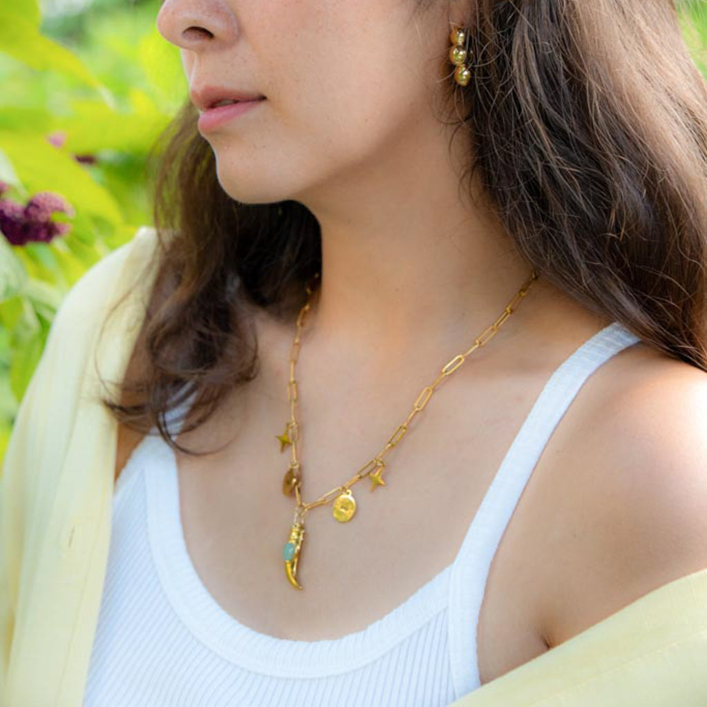 A close-up lifestyle shot of a woman wearing a delicate gold chain necklace with a crescent moon and star pendant, styled with a white ribbed tank top and a light yellow cardigan, set against a soft-focus green garden background.