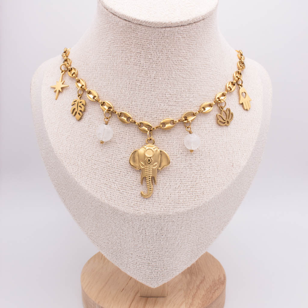 A gold-toned charm necklace displayed on a beige fabric jewelry bust. The necklace features an elephant pendant, a star charm, leaf and palm leaf charms, and two small white bead-like accents.