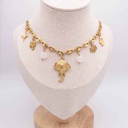 A gold-toned charm necklace displayed on a beige fabric jewelry bust. The necklace features an elephant pendant, a star charm, leaf and palm leaf charms, and two small white bead-like accents.