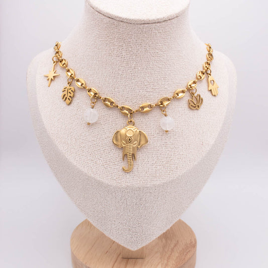 A gold-toned charm necklace displayed on a beige fabric jewelry bust. The necklace features an elephant pendant, a star charm, leaf and palm leaf charms, and two small white bead-like accents.