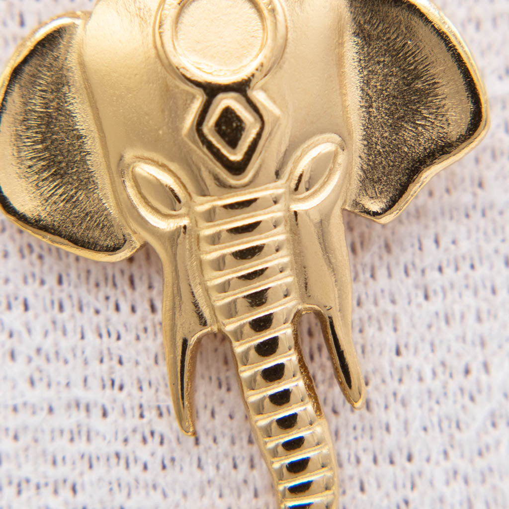 A close-up of a gold-toned elephant head brooch or pin with textured ears, a patterned forehead, and a trunk, shown on a white textured fabric background.