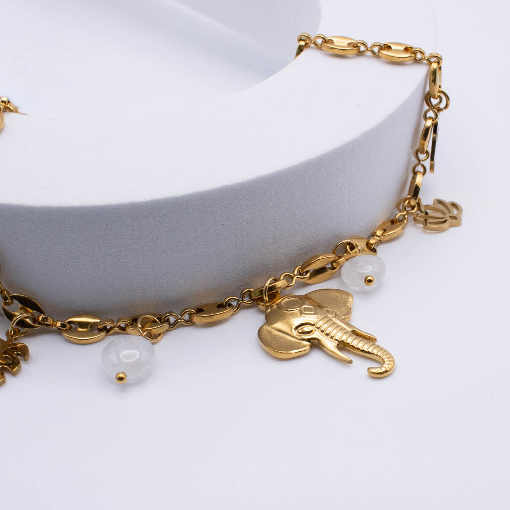 Gold chain bracelet with an elephant charm, white pearl beads, and a small gold monogram pendant, displayed on a white curved stand.