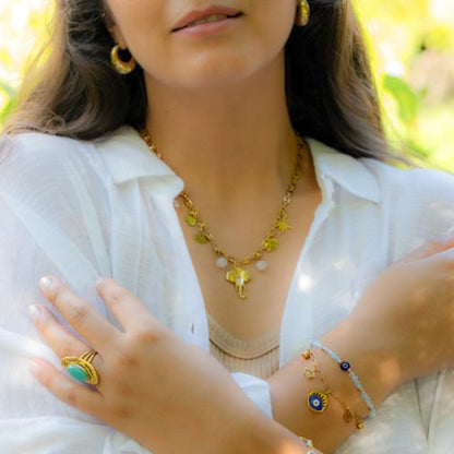 A close-up lifestyle shot of a woman wearing a gold necklace with animal charms, a matching gold ring with a turquoise stone, and delicate gold bracelets on her wrist, set against a soft, blurred green background.
