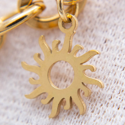 A close-up of a gold-toned sunburst charm with a cutout center, attached to a matching chain, resting on a textured off-white fabric background.