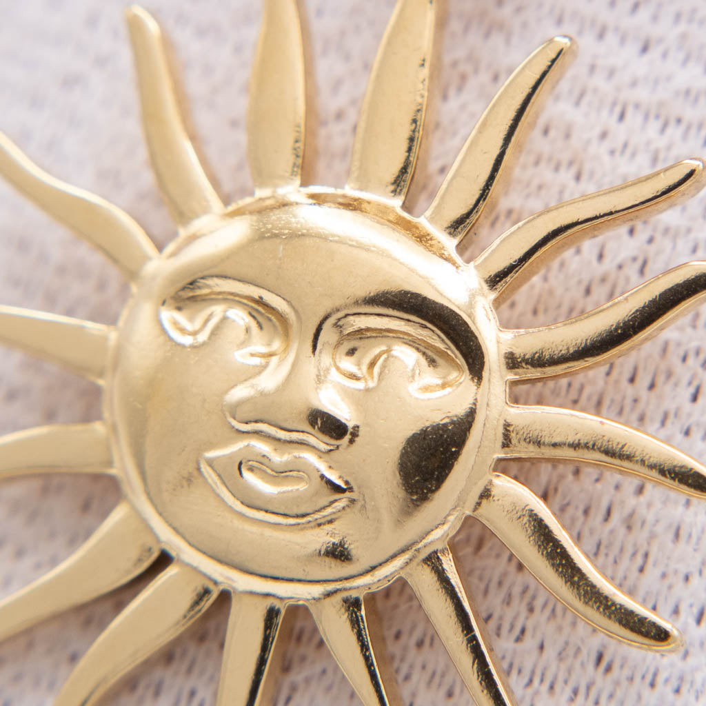 A close-up of a gold-toned sun face charm or brooch with radiating rays, shown against a textured off-white fabric background.