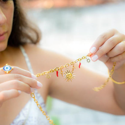 A close-up lifestyle shot of a woman holding a delicate gold chain bracelet with sun and star charms, wearing a matching gold chain bracelet on her wrist and a blue and white evil eye ring on her finger.