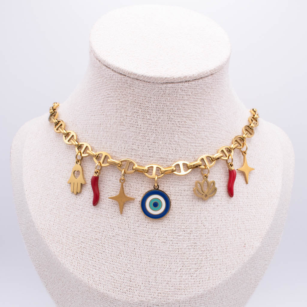 Gold-tone charm necklace displayed on a cream fabric jewelry bust. The necklace features a heart-link chain with multiple dangling charms, including a blue and white evil eye, gold stars, a hamsa hand, a lotus flower, and small red italian horn.