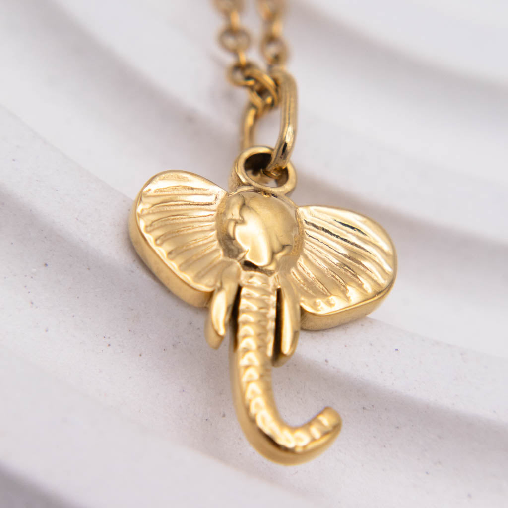 A gold elephant pendant necklace with detailed textured ears and trunk, hanging from a gold chain against a white textured background.