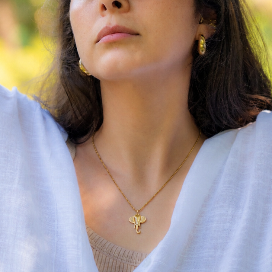 A woman outdoors wearing gold jewelry, including a gold necklace with a small cross pendant, rings, and bracelets with turquoise and blue accents, while shielding her face from the sun.