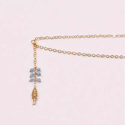A delicate gold chain necklace with a lariat-style drop featuring light blue gemstone beads and a textured gold pendant, displayed on a soft pink background.