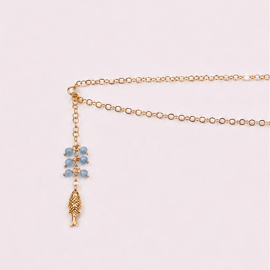 A delicate gold chain necklace with a lariat-style drop featuring light blue gemstone beads and a textured gold pendant, displayed on a soft pink background.