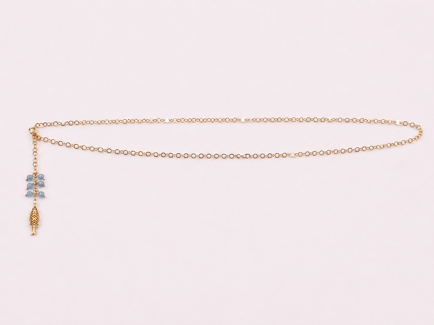 A delicate gold chain necklace with a small blue and gold charm, displayed on a light pink background.
