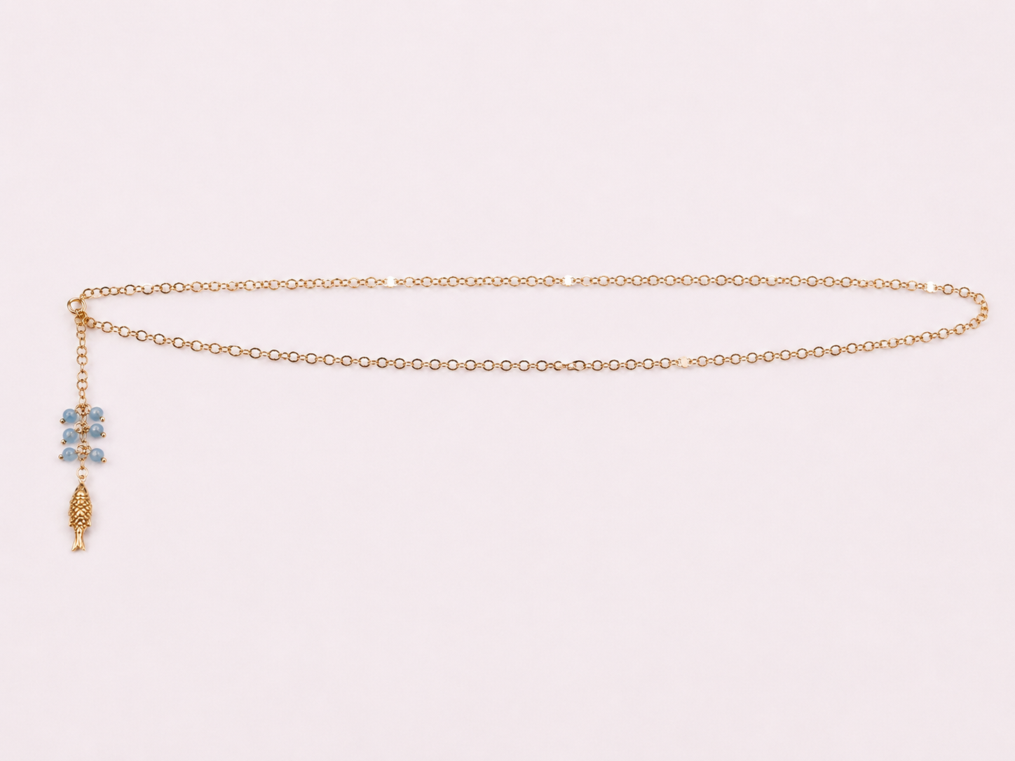 A delicate gold chain necklace with a small blue and gold charm, displayed on a light pink background.