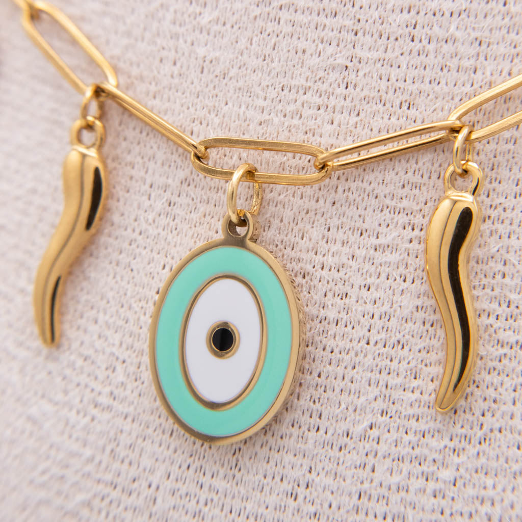Gold-tone chain necklace with an oval evil eye pendant in mint green, white, and black, flanked by two small wavy gold charms, displayed on a textured cream fabric.