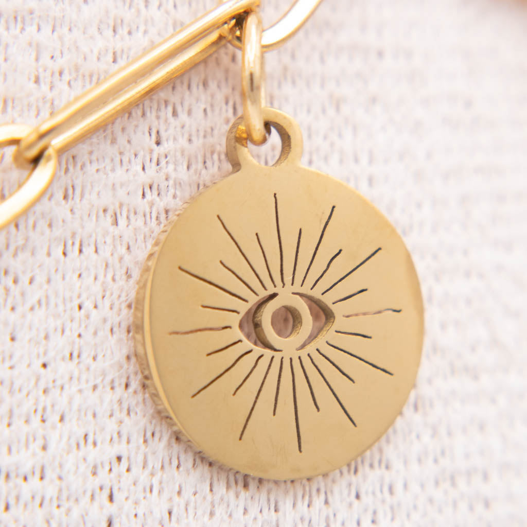 A close-up of a round gold-toned pendant charm with an engraved eye and radiating lines, attached to a gold chain against a textured off-white fabric background.