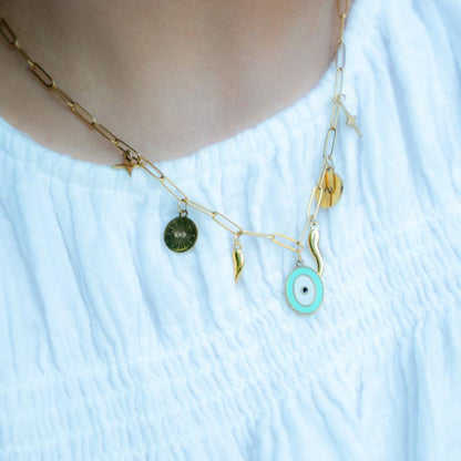 A close-up of a gold chain necklace with multiple charms, including a mint green evil eye pendant and a dark green sun disc, worn by a person in a white textured top.