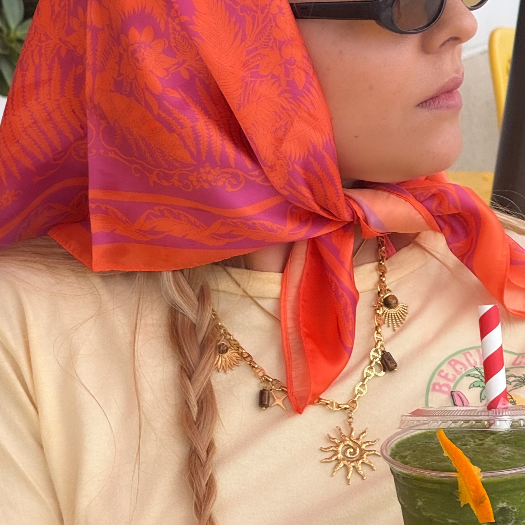 Woman wearing a red scarf and sunglasses, holding a green smoothie with a straw.