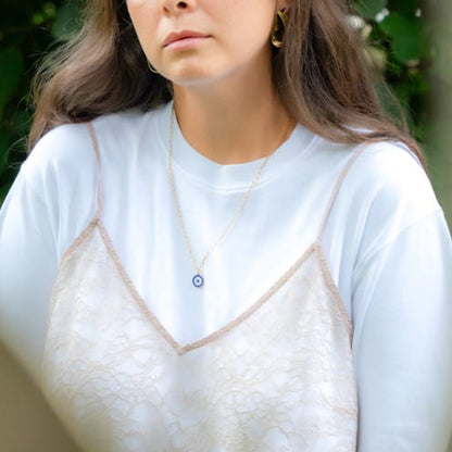 A close-up of a woman wearing a delicate gold necklace with a blue and white evil eye pendant, layered over a white long-sleeve shirt and a white lace camisole.