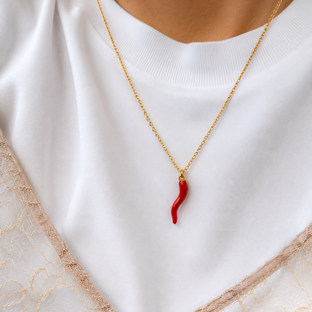 A close-up of a gold chain necklace with a small, wavy red pendant, worn by a person in a white ribbed top and beige lace.