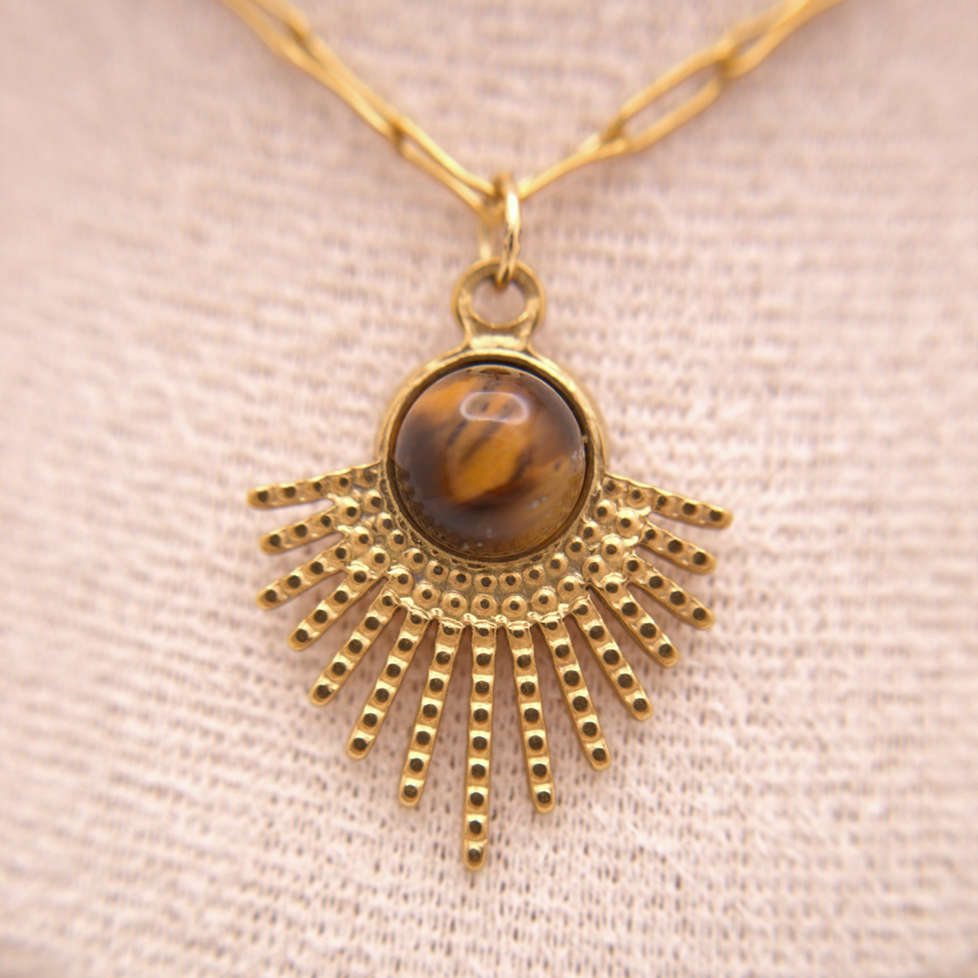 Gold necklace with a sunburst pendant featuring a tiger's eye stone on a beige fabric background