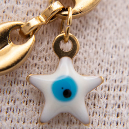 Gold-tone metal keychain with a white star-shaped evil eye charm featuring a blue and black center, shown on a textured beige fabric background.
