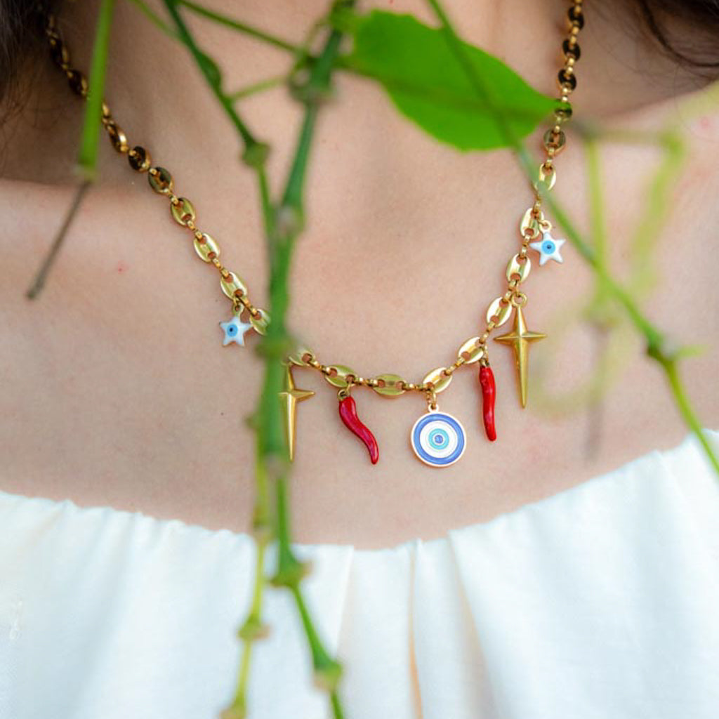 A gold-toned necklace worn on a person's neck, featuring small star charms, lightning bolts, red chili pepper charms, and an evil eye target charm, with blurred green leaves in the foreground.