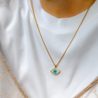 A gold chain necklace with a small evil eye pendant featuring a turquoise blue iris, worn by a person in a white top and beige lace garment.