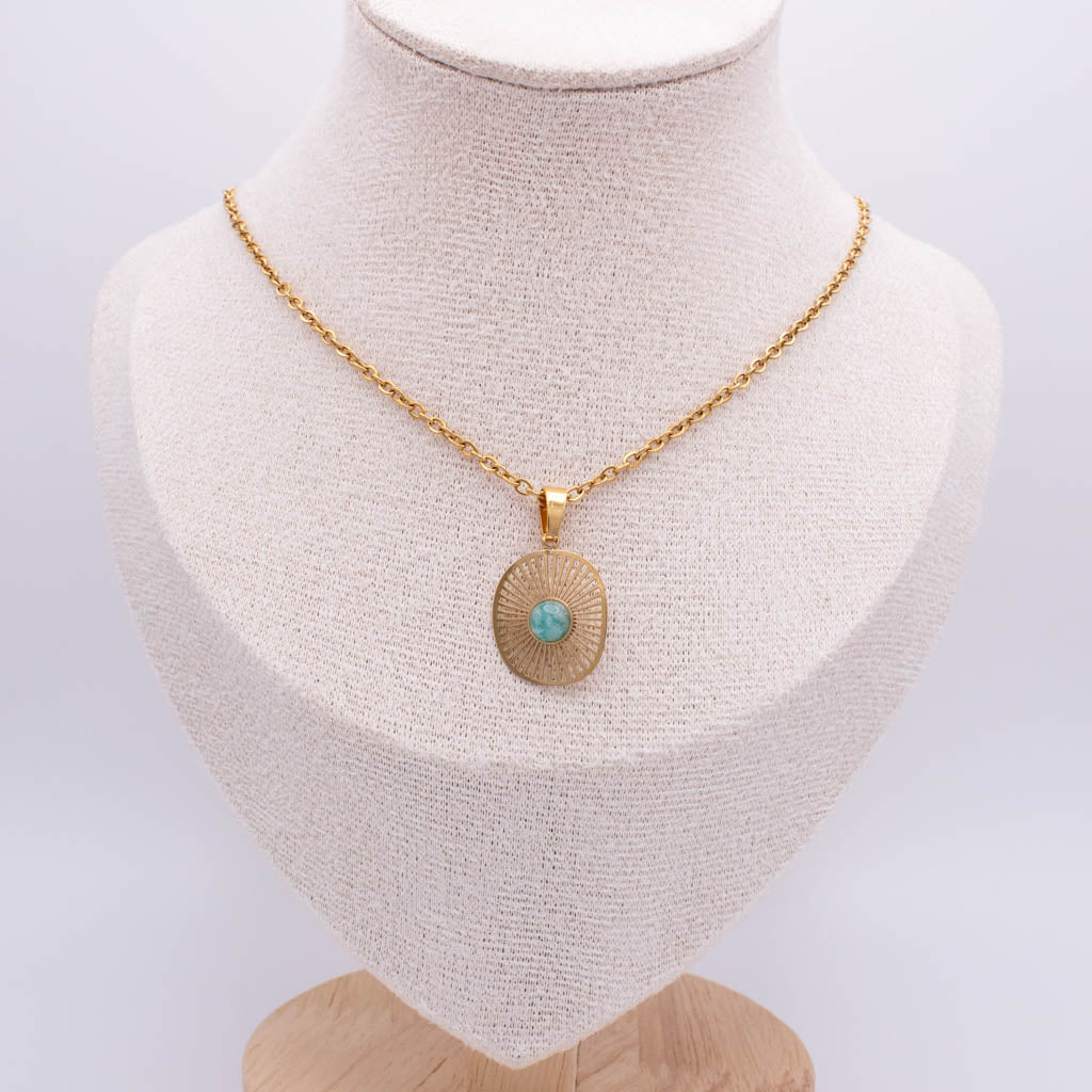 A gold chain necklace with a round sunburst pendant featuring a small turquoise stone in the center, displayed on a cream-colored fabric jewelry bust.