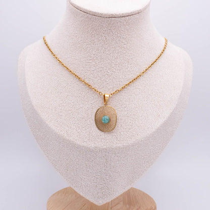 A gold chain necklace with a round sunburst pendant featuring a small turquoise stone in the center, displayed on a cream-colored fabric jewelry bust.