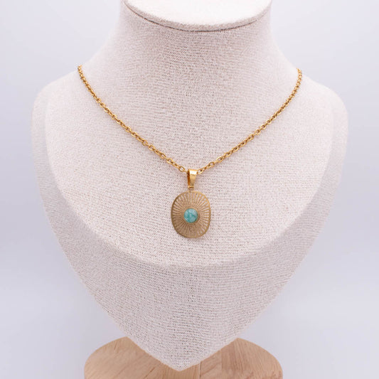 A gold chain necklace with a round sunburst pendant featuring a small turquoise stone in the center, displayed on a cream-colored fabric jewelry bust.
