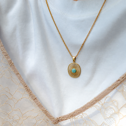 A gold pendant necklace with a circular sunburst design and a small turquoise center, worn by a person in a white top and beige lace garment.